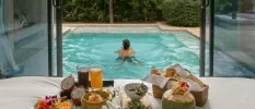 Man swims in a pool in front of a bedroom laid up for breakfast with fresh fruit, coconut juice, a basket of pastries and fresh juices