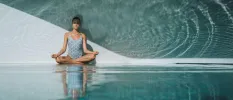 Woman in blue patterned swimsuit sits cross legged with her eyes closed next to a pool with a wave mural on the wall next to her