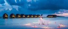 Staff member in white lights a lantern on a table set on the beach, with lanterns all around and overwater villas in the background