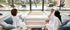 Three woman in white robes sit by an indoor pool with floor-to-ceiling windows overlooking an outdoor pool and gardens