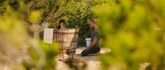 Female sits in a wooden bathtub as a male sits on his knees beside her in a leafy garden