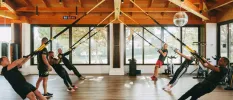 Group in active wear take part in a TRX class with ropes hanging from a wood-beamed ceiling in a studio with floor-to-ceiling windows