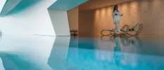 Woman in white walks past an indoor pool past a wall with an art sculpture and softly lit by spotlights