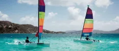 Two people sailing in the ocean with multi-coloured sails with a green island in the distance