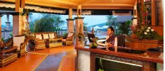 Receptionist in authentic Thai dress stands behind a desk in a traditionally designed reception area, with tiled floors, rattan chairs, dark-wood panelling and a view to the hillsides outside