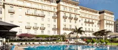 Palacio Estoril's grand, 1930s facade with white walls, black wrought-iron balconies and a swimming pool under a blue sky