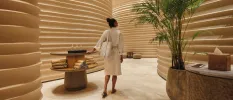 White-robed woman walking into a spa corridor with curved walls, a potted plant and a table stacked with towels and clear bottles