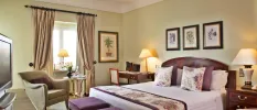 Charming, homey hotel room with white bedding with purple floral cushions and throw, a cosy beige armchair with a purple cushion, beige curtains and floral pictures on the walls