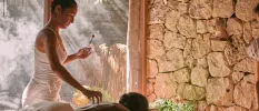 Therapist in white carries out a spa treatment as a woman lies on her front in a room with a cobbled stone wall