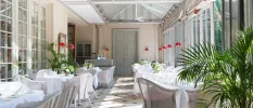 Tables with crisp white tablecloths lined up in sunny conservatory area, with green plants, a patterned floor and red hanging lights