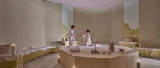 White-robed woman has her arm massaged by a therapist in a candlelit marble-cloaked room with cream walls and floors