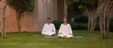 Man and woman in white meditate on mats in a lawned garden