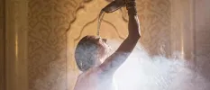 Woman pouring water over her head as steam rises around her in front of a Moroccan-style wall