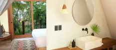 Airy bedroom and bathroom with a patterned rug, full-length balcony doors with a view of greenery, a wooden vanity unit and a white butler sink, plus a round mirror on the wall
