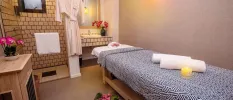 Treatment room with a cream tiled shower and vanity unit, a wooden side table, and massage bed with a blue blanket