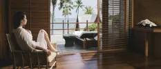 White-robed women sits in a hotel room decked out in dark woods, overlooking a furnished balcony and the palm trees and ocean in the distance