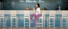 Smiling woman in purple leggings and a white T shirt sits at a bar sipping an orange smoothie or juice, with a sign behind her that reads 'The Vitamin Bar'