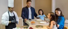 Man, woman and two daughters smile as they sit around a table with a chef on the other side and food spread in front of them