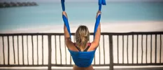 Woman in active wear hangs from blue ribbon on a terrace overlooking a white beach and turquoise shallows