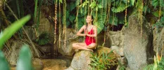 Woman in a red swimsuit sits on a rock overlooking a pond with her hands clasped in prayer and smiling