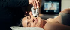 Smiling female lies on her back on a massage bed as a therapist uses a device on her face, with a medical-style digital screen in the background