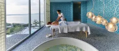 Therapist massages a woman on a bed in a room of blue mosaic tiles, with a round bath filled with white flowers and a large window with sea views