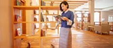 Woman in traditional dress reading a book next to a library shelf in a room with a wooden floor and wooden beams 