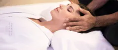 White-robed woman lies on a bed with her eyes closed as a therapist massages her head