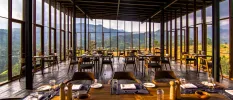 Restaurant with panoramic glass windows, overlooking mountains and greenery, designed in dark woods and with tables laid up for dining