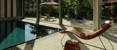 Indoor pool next to a wooden lounger with a white towel, and a wooden table with a glass of water, all overlooks a sunny terrace through full-length windows