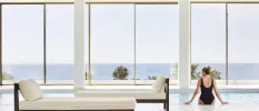 Woman in a black swimsuit sits at the pool edge inside a spa, with white-cushioned loungers behind her and full-length windows with a sea view