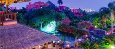 Lit red buildings with thatched rooftops, an outdoor terrace with tables and chairs and tropical greenery, with the Alicante skyline in the background
