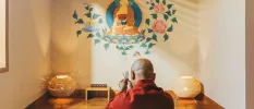 Bald man in red sews in a cosy room with a colourful floral and Buddha mural on the wall, and two rattan lantern-style floor lamps