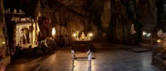 Two staff members in white stand at the centre of a cave-like space, with candlelight, statues and a shrine to the left-hand side