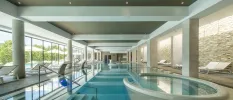 Indoor swimming pool with built in whirlpool, surrounded by white loungers and with floor-to-ceiling windows