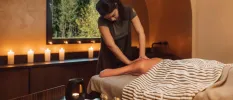 Woman lies on her front and enjoys a massage in a candlelit room, with a dark-wood vanity and window overlooking the forest