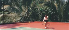 Tennis court with jungle surroundings and a player in sunglasses about to hit the ball