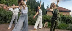 Four woman in loose fitting clothes stretch their arms and legs out on an outdoor terrace surrounded by gardens