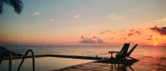 Corner of an infinity pool with wooden decking overlooking the sea under a sunset-filled sky
