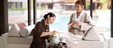 Staff member in brown pours tea for a woman in a robe sitting on a daybed in front of a pool