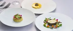 White plates with small portions of intricately presented food, including one designed to look like a yellow flower, and another with tiny vegetables arranged in a circle around some fish