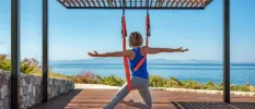 Woman in active wear hanging in an an aerobic harness in a yoga pose, on a wooden terrace with a view of the Aegean Sea
