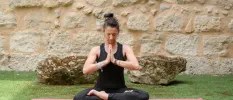 Woman sitting with her legs crossed and hands clasped in prayer wearing black active wear on a pink yoga mat in a stone-walled courtyard