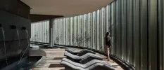 A woman in a black swimsuit walks past floor-to-ceiling glass window panels in a room with curved loungers and a shower area with a foot pool