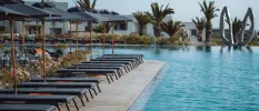Swimming pool under a blue sky, edged by palm trees, grey parasols and grey-cushioned loungers, and with a sculpture of donkey ears in the background