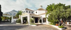 Marbella Club entrance, with a paved driveway, low-rise whitewashed buildings with terracotta roof tiles and surrounding greenery, with mountains in the background