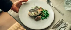 Waiter lays down a white plate of grilled fish, peas and shallot