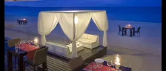 Wooden terrace with tables lit by lanterns, with the beach in front and a four-poster cabana with white drapes