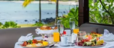 Table laid for breakfast, with two plates of eggs, beans, tomato and bacon, a basket of bread, a bowl of fruit and two glasses of orange juice, with the ocean in the background