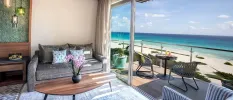 Hotel room seating area with a grey sofa, blue rug and wooden coffee table, next to an open furnished balcony overlooking the beach